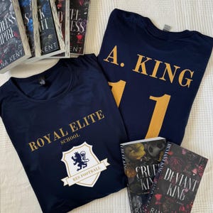 Puede incluir: Dos camisetas azul marino con letras doradas. Una camiseta dice "Royal Elite School" con un escudo y la otra dice "A. King 11". Tres libros con los títulos "Deviant King" y "Cruel King" son visibles en el fondo.