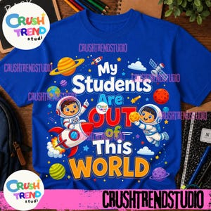 May include: A royal blue t-shirt featuring a space-themed graphic with two cartoon astronauts, a rocket ship, planets, stars, and clouds. The text reads My Students Are OUT of This WORLD in a mix of white, red, and yellow bold lettering.