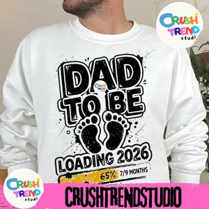 May include: White sweatshirt with a black and white graphic that reads "DAD TO BE LOADING 2026". The design includes baby footprints and the text "65% 7/9 MONTHS". The sweatshirt has a crew neck.