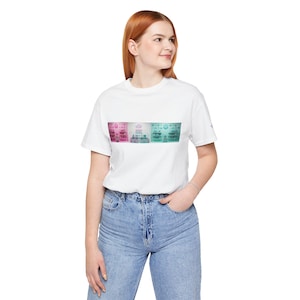 Puede incluir: Una camiseta blanca de manga corta con un estampado gráfico rectangular en el pecho. El gráfico muestra una serie de estantes llenos de diversos artículos, con una transición de color que va del rosa a la izquierda al turquesa a la derecha.