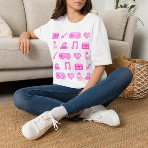 Puede incluir: Una camiseta blanca de manga corta con un patrón de cuadrícula repetitivo de iconos de arte de píxeles rosas, que incluyen mandos de juego, corazones, cofres, espadas, sombreros de mago, notas musicales y pociones, con vaqueros azul oscuro.