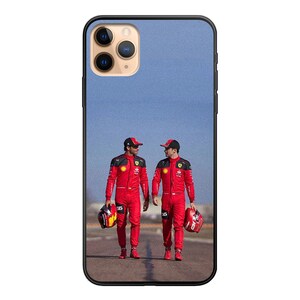 May include: Two people wearing red racing suits and helmets stand on a track. They are holding helmets in their hands. The background is a blue sky.