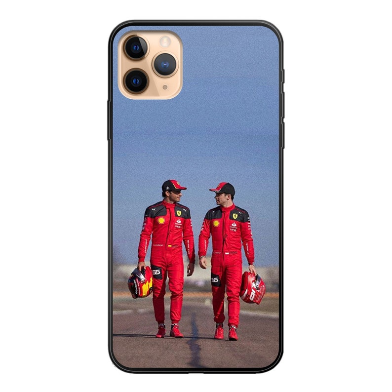 May include: Two people wearing red racing suits and helmets stand on a track. They are holding helmets in their hands. The background is a blue sky.