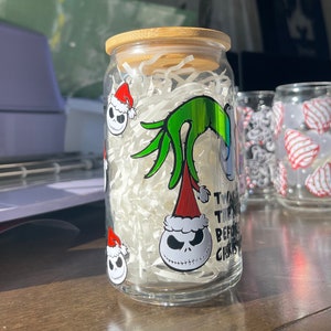Puede incluir: Un tarro de vidrio transparente con tapa de bamb&uacute;, decorado con pegatinas de tem&aacute;tica navide&ntilde;a con el Grinch y Jack Skellington. El tarro est&aacute; lleno de cacahuetes de embalaje blancos. El texto en el tarro dice "Twas the night before Christmas".