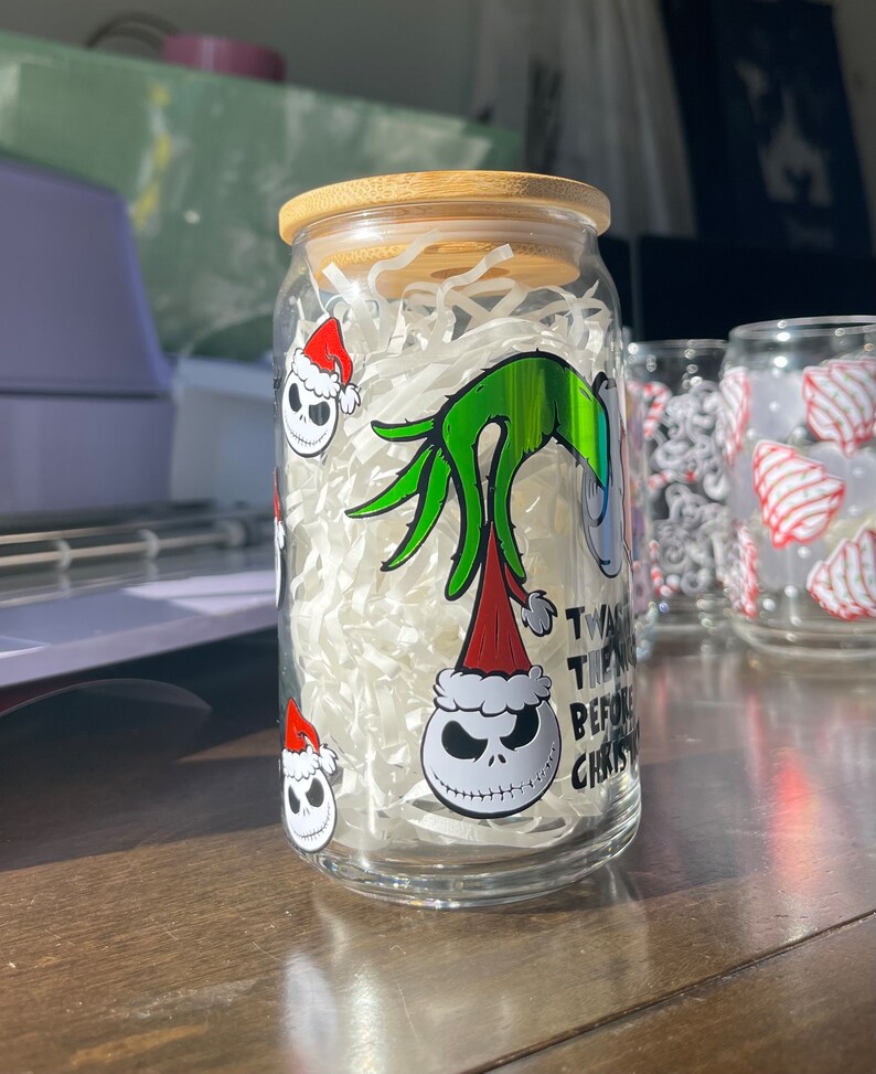 Puede incluir: Un tarro de vidrio transparente con tapa de bamb&uacute;, decorado con pegatinas de tem&aacute;tica navide&ntilde;a con el Grinch y Jack Skellington. El tarro est&aacute; lleno de cacahuetes de embalaje blancos. El texto en el tarro dice "Twas the night before Christmas".