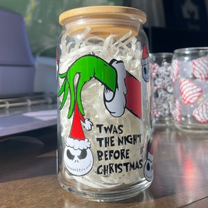 Puede incluir: Un tarro de vidrio transparente con tapa de madera, decorado con una mano verde que sostiene un gorro de Pap&aacute; Noel rojo, y una cara de Jack Skellington en blanco y negro. El tarro tiene el texto "Twas the night before Christmas" impreso en &eacute;l.