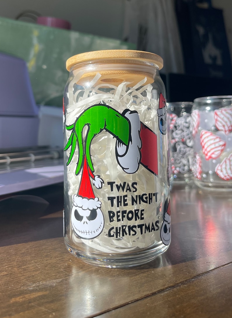 Puede incluir: Un tarro de vidrio transparente con tapa de madera, decorado con una mano verde que sostiene un gorro de Pap&aacute; Noel rojo, y una cara de Jack Skellington en blanco y negro. El tarro tiene el texto "Twas the night before Christmas" impreso en &eacute;l.