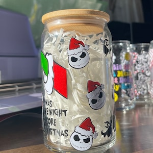 Puede incluir: Un tarro de vidrio transparente con tapa de bamb&uacute; lleno de papel blanco triturado. El tarro tiene una variedad de pegatinas, incluyendo un personaje con un sombrero verde, un personaje con un sombrero rojo y el texto "Twas the Night Before Christmas".