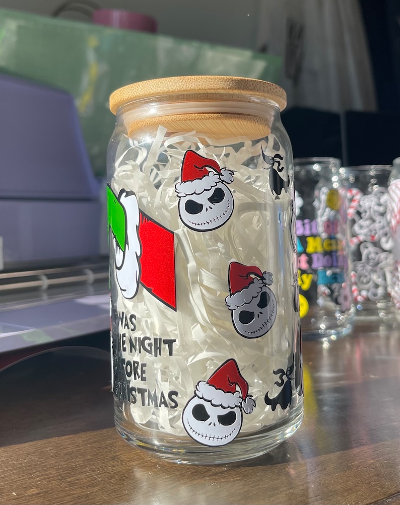 Puede incluir: Un tarro de vidrio transparente con tapa de bamb&uacute; lleno de papel blanco triturado. El tarro tiene una variedad de pegatinas, incluyendo un personaje con un sombrero verde, un personaje con un sombrero rojo y el texto "Twas the Night Before Christmas".