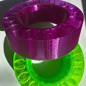 May include: Two 3D printed rings, one purple and one green, with a decorative pattern around the edge. The rings are stacked on top of each other.