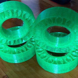 May include: Four bright green, 3D-printed circular objects with a repeating pattern of small, rounded indentations. The objects are stacked on top of each other, with a wooden surface in the background. The objects have a hollow center.