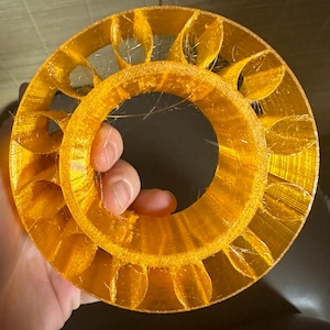 May include: A 3D printed, orange, circular object with a hollow center and a series of evenly spaced, curved, outward-facing blades.