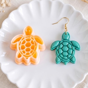 May include: A peach-colored plastic turtle-shaped cookie cutter and a matching teal turtle-shaped earring with a gold-toned metal hook. Both items feature a detailed shell pattern with a central hexagonal design and are displayed on a white plate.