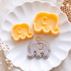 May include: Two yellow plastic elephant-shaped cookie cutters of different sizes and one grey speckled clay elephant pendant with a gold-colored metal jump ring attached to the top, all arranged on a white scalloped plate.