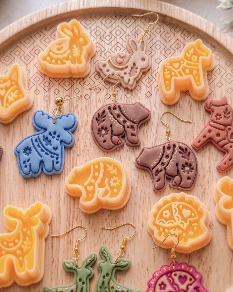 Scandi folk animal clay cutters displayed with finished polymer clay earrings on a wooden tray, featuring deer, moose, bunny and bear designs