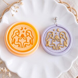 Op de afbeelding: Een ronde oranje plastic koekjesvorm met een Tweelingen sterrenbeeldontwerp met twee figuren, sterren en een maansikkel. Daarnaast een bijpassende ronde hanger van lavendelkleurige klei met een goudkleurig reliëf van het Tweelingen-teken.