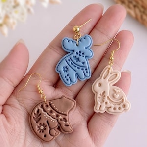 Close-up of handmade polymer clay earrings in hand, featuring moose, bunny and bear shapes with detailed imprint patterns