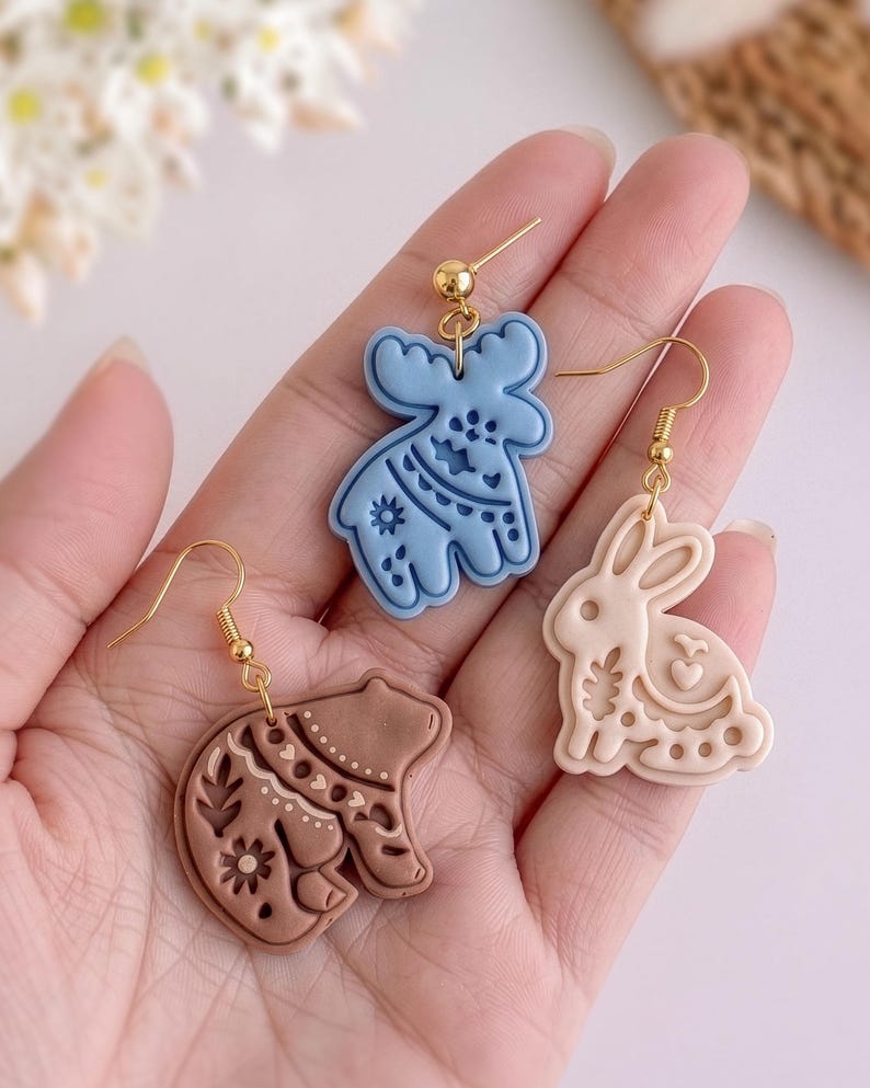 Close-up of handmade polymer clay earrings in hand, featuring moose, bunny and bear shapes with detailed imprint patterns