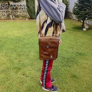 May include: A brown leather crossbody bag with a flap closure and a metal buckle. The bag is worn over a red and black patterned pants with white accents.