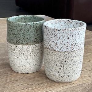 May include: Two ceramic tumblers with a speckled design. One tumbler has a sage green top and a speckled white bottom. The other tumbler has a speckled white top and bottom. The tumblers are sitting on a wooden surface.