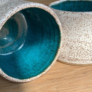 May include: Two ceramic mugs with a speckled white exterior and a vibrant turquoise interior. The mugs are set on a wooden surface, showcasing their contrasting colors and textures. The design suggests a handcrafted or artisanal aesthetic.