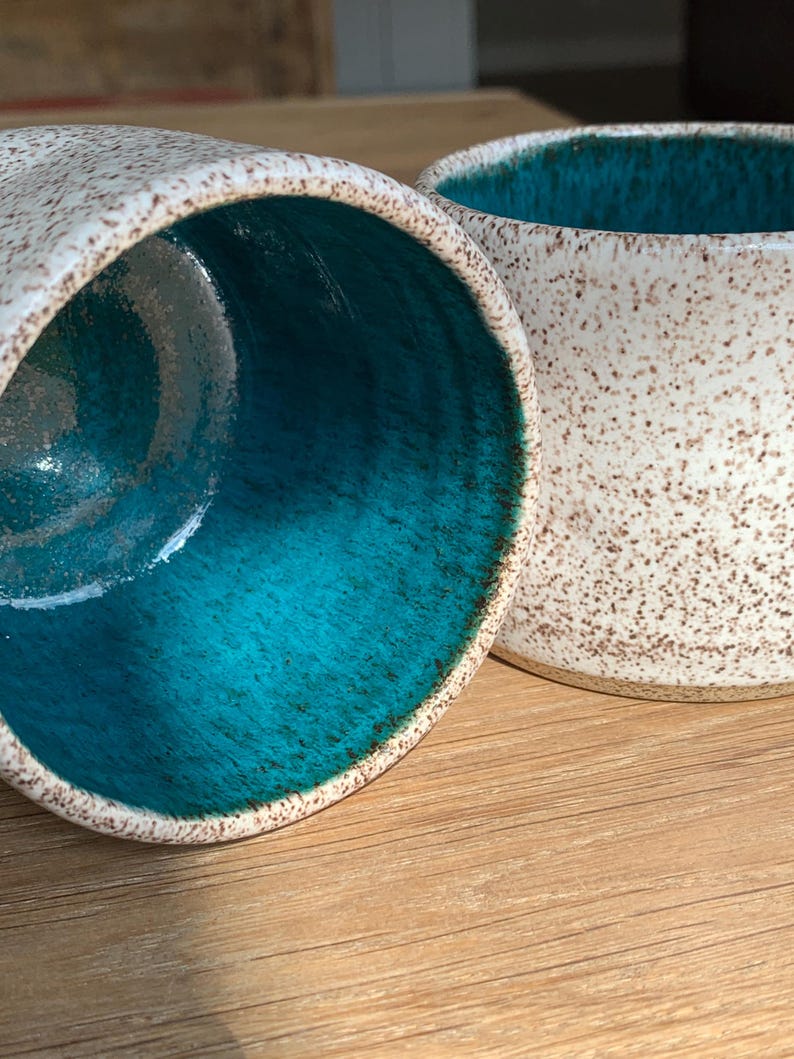 May include: Two ceramic mugs with a speckled white exterior and a vibrant turquoise interior. The mugs are set on a wooden surface, showcasing their contrasting colors and textures. The design suggests a handcrafted or artisanal aesthetic.