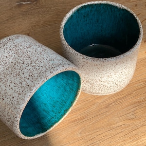 May include: Two ceramic cups with a speckled white exterior and a vibrant teal interior. One cup is upright, the other is on its side, both showcasing the contrasting colors. The cups are set on a wooden surface.