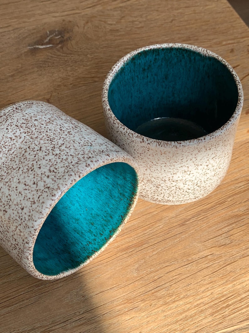May include: Two ceramic cups with a speckled white exterior and a vibrant teal interior. One cup is upright, the other is on its side, both showcasing the contrasting colors. The cups are set on a wooden surface.