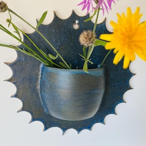 May include: A dark blue ceramic wall planter with a sunburst-shaped backplate and a rounded pocket for flowers. The piece features a glossy, mottled glaze with deep indigo and charcoal tones, and includes a small hole at the top for mounting.