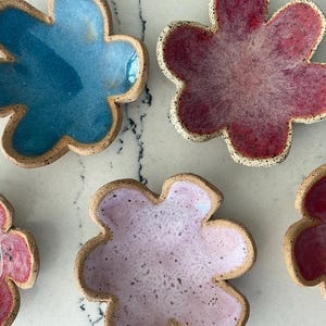 May include: A collection of handmade ceramic flower-shaped dishes in various colors. The dishes feature a speckled, glazed interior in shades of blue, pink, and lavender, with a natural, unglazed rim. These decorative dishes are ideal for serving small portions.