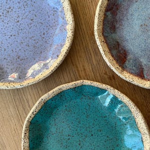 May include: Three handmade ceramic plates in shades of blue and teal. Each plate has a speckled, textured surface and a light-colored rim. The plates are arranged on a wooden surface, showcasing their unique colors and textures.