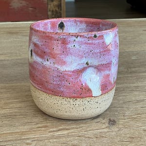 May include: A ceramic tumbler featuring a two-tone design with a speckled beige base and a glossy, mottled pink and white upper section. The cylindrical vessel has a smooth, rounded rim and a slightly tapered shape, showcasing a handcrafted aesthetic.