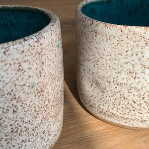 May include: Two ceramic mugs with a speckled, off-white exterior and a teal interior. The mugs have a cylindrical shape and are likely handmade. The speckled pattern is brown. The mugs are placed side by side.