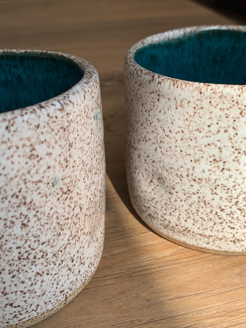 May include: Two ceramic mugs with a speckled, off-white exterior and a teal interior. The mugs have a cylindrical shape and are likely handmade. The speckled pattern is brown. The mugs are placed side by side.