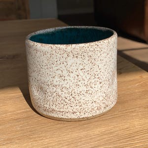 May include: A speckled ceramic mug with a teal interior. The exterior is off-white with brown speckles. The mug is cylindrical and sits on a wooden surface, catching the sunlight.