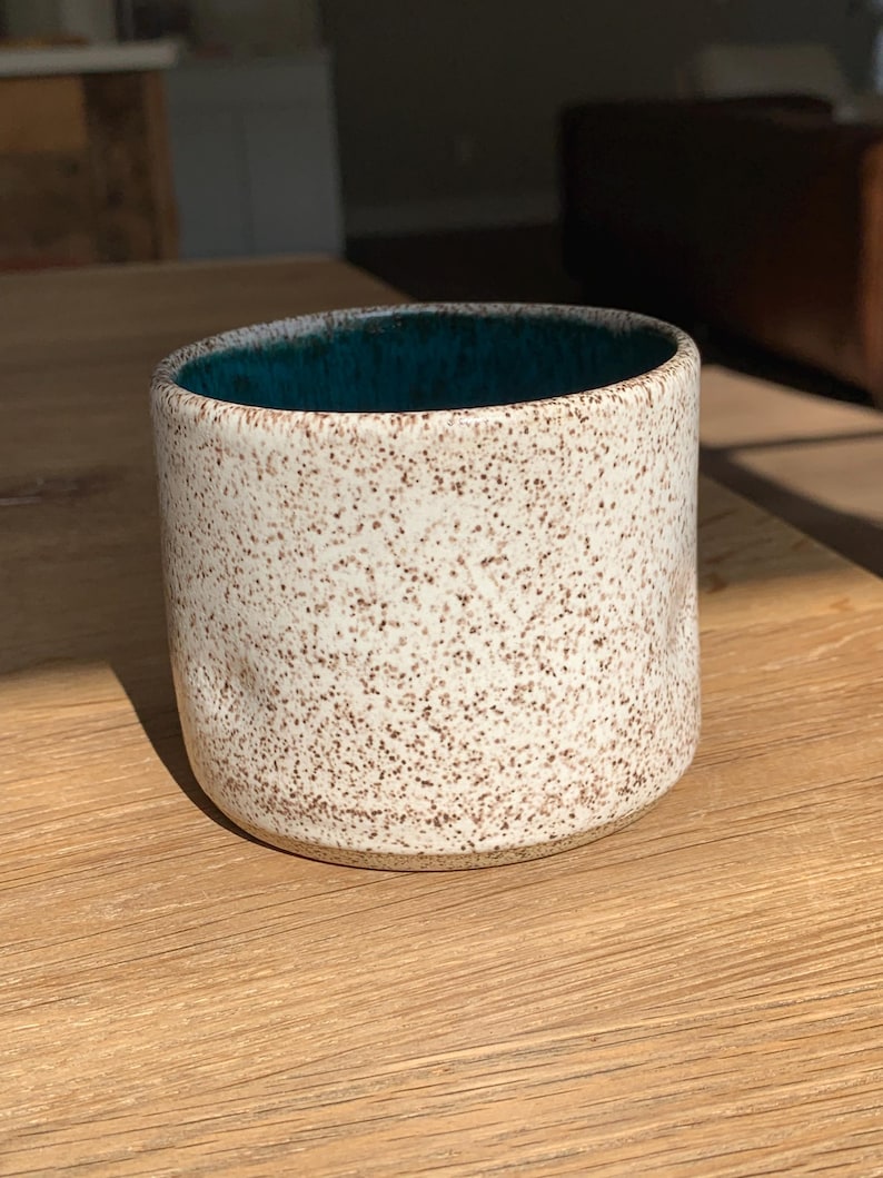 May include: A speckled ceramic mug with a teal interior. The exterior is off-white with brown speckles. The mug is cylindrical and sits on a wooden surface, catching the sunlight.