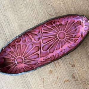 May include: An oval ceramic tray with a dark, textured charcoal-colored rim and a vibrant, glossy magenta interior. The surface features two embossed, symmetrical floral patterns with radiating petals that catch the light.