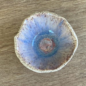 May include: A small, handmade ceramic bowl with a unique, organic shape. The interior features a blend of blue and purple hues, with a speckled, textured rim. The center has a reddish-brown accent. A decorative piece for the home.