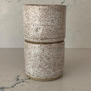 May include: Two stacked ceramic cups with a speckled white glaze and brown flecks. The cups have a cylindrical shape and a natural, earthy aesthetic. The rims and bases are unglazed, revealing the natural clay color.