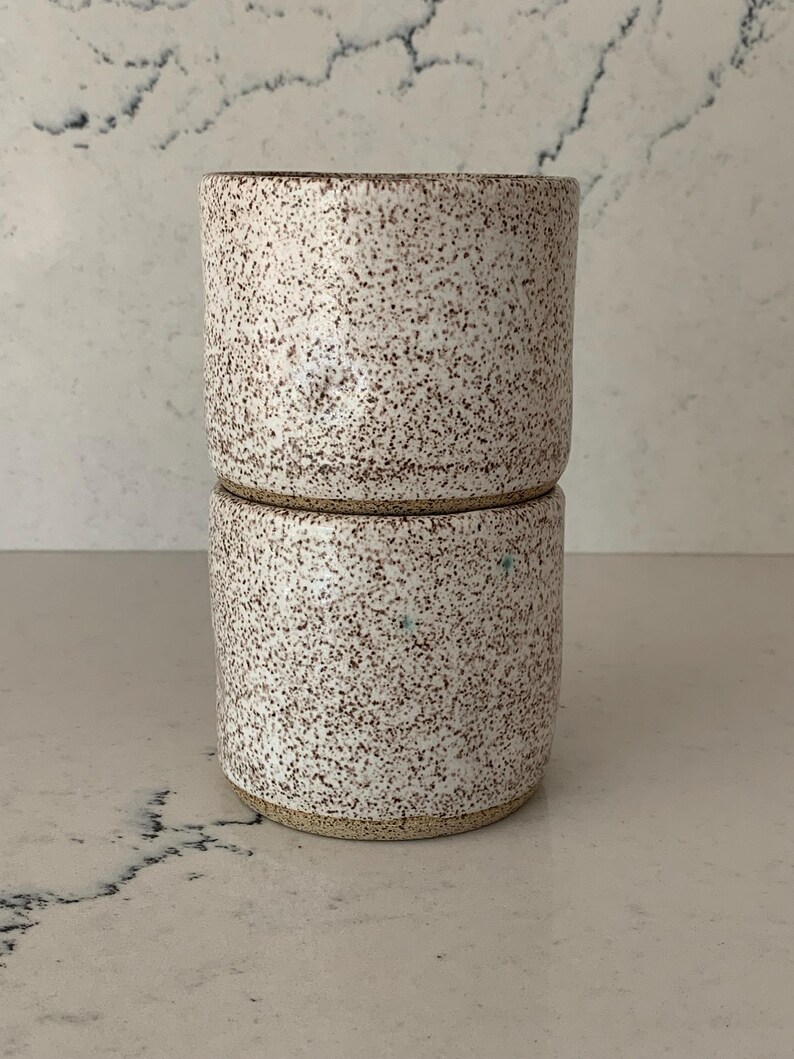 May include: Two stacked ceramic cups with a speckled white glaze and brown flecks. The cups have a cylindrical shape and a natural, earthy aesthetic. The rims and bases are unglazed, revealing the natural clay color.