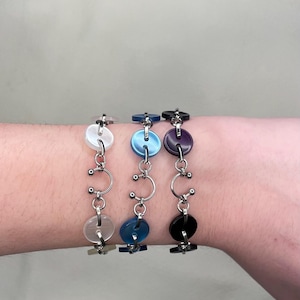 Puede incluir: Tres pulseras de botón plateadas con botones de diferentes colores. Las pulseras tienen una cadena plateada con un diseño circular. Los botones son blancos, azules y morados.