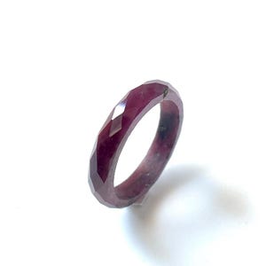 May include: A faceted, deep red ring. The ring has a geometric design with multiple facets that catch the light. The ring is set against a white background, highlighting its color and shape. The ring appears to be made of a gemstone.