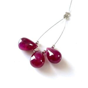 May include: A necklace featuring three teardrop-shaped, deep red gemstones. The gems are strung on a thin, silver-colored wire, with small silver beads separating them. The background is plain white, highlighting the jewelry.