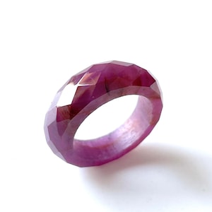 May include: A faceted, ruby-colored ring. The ring is a deep pinkish-red hue, with multiple facets that catch the light. The gemstone ring is set against a white background.