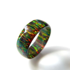 Può includere: Un anello a fascia lucido con una base scura e vivaci macchie iridescenti di verde, arancione e rosso su tutta la superficie. Il materiale presenta una finitura lucida che riflette la luce, creando un aspetto multicolore e cangiante sull'intera struttura circolare dell'anello.