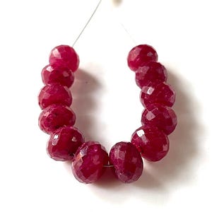 May include: Strung faceted ruby beads. The beads are a deep red color and have a multi-faceted surface. The beads are strung on a thin, clear wire, and are displayed against a white background. The beads are likely for jewelry making.
