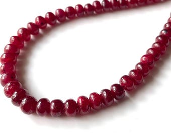 May include: A strand of deep red, round gemstone beads. The beads are strung together on a thin, clear string, and are evenly sized. The beads have a polished, glossy finish and are set against a white background.