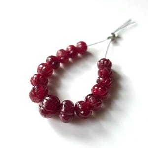 May include: A strand of deep red, carved gemstone beads. The beads are strung on a thin, light blue string and are arranged in a curved shape. The beads have a glossy finish and are likely used for jewelry making.