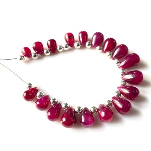 Puede incluir: Un collar de cuentas de rubí pulidas en forma de lágrima. Las cuentas son de un color rojo intenso y rico y están ensartadas con pequeñas cuentas espaciadoras plateadas. Las cuentas se muestran sobre un fondo blanco.