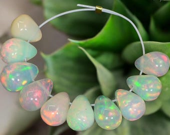May include: A strand of pear-shaped opal beads. The opals display a milky white base color with flashes of iridescent colors, including pink, green, and orange. The beads are strung on a white cord, with a gold-colored bead at the top.